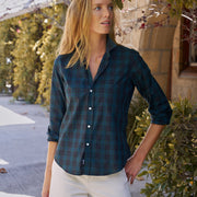 A woman wearing a dark teal and green plaid button-down shirt with white pants, standing outdoors in front of a stone wall and green foliage.
