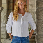 A woman with long blonde hair wearing a white long-sleeve button-up shirt and blue jeans, standing in front of a stone column.