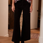 A person wearing black wide-leg pants with a red, white, and black plaid button-up shirt and black pointed-toe heeled boots, standing in a home interior with wood floors.