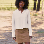 A woman with curly black hair stands outdoors wearing a cream long-sleeve cropped button-front top and a brown mini skirt, with a blurred natural background of trees and grass.