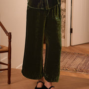 A woman stands indoors wearing a dark green velvet matching lounge set with a long-sleeve button-front top and wide-leg pants. The room has light wood flooring, patterned wallpaper, a chair with a candle, and a wall sconce.