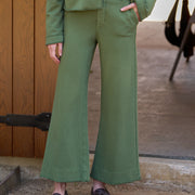 A person wearing an olive green tailored pantsuit with a cropped jacket and wide-leg trousers, paired with a white top and dark brown loafers, standing outdoors against a wooden door and concrete ground.
