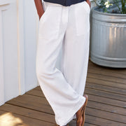 A woman stands on a wooden deck wearing a dark navy long-sleeve button-up shirt and white wide-leg pants, with brown sandals. White house siding, a window, and a large galvanized planter with climbing white flowers are in the background.