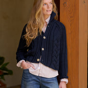 A woman wearing a navy blue cable-knit button-front cardigan over a striped shirt and blue jeans, standing indoors next to a wooden door.