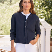 Woman wearing a navy cardigan over a white shirt and pants, standing outdoors with greenery in the background.