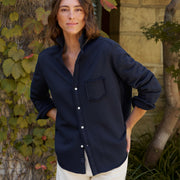 A woman wearing a dark navy blue long-sleeve button-up shirt with a chest pocket, paired with off-white pants, standing outdoors in front of a stone wall and leafy vines.