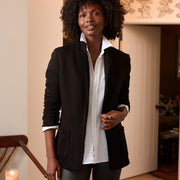 A woman with curly dark hair stands indoors wearing a black blazer over a white button-up shirt and dark gray trousers. The room has warm beige walls, a wooden chair with a lit candle, and a doorway in the background.