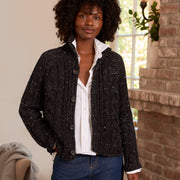 A woman with curly black hair stands indoors wearing a black sequin knit cardigan with a white button-up shirt and blue jeans. The setting features a warm brick fireplace on the right and a neutral wall on the left.