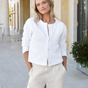A woman wearing a white long-sleeve button-front cardigan and light beige linen trousers, standing in a sunny outdoor corridor with columns.