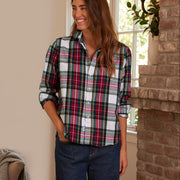 A woman wearing a long-sleeve button-up plaid shirt with red, green, black, and white stripes, paired with dark blue jeans, standing in a cozy room with a brick fireplace and window.