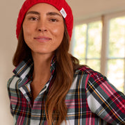 A woman wearing a bright red ribbed knit beanie hat and a red, black, and white plaid button-up shirt, photographed in a softly lit indoor setting.