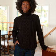 A woman with curly black hair wearing a black cable-knit turtleneck sweater and blue plaid pants, standing in a bright, modern home interior.