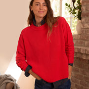 A woman wearing a bright red ribbed knit sweater over a dark navy collared shirt, paired with dark blue jeans, standing in a warmly lit room with a brick pillar and window in the background.
