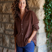 A woman with curly brown hair wearing a brown corduroy button-down shirt and blue jeans, standing against a stone wall with green ivy on the right.