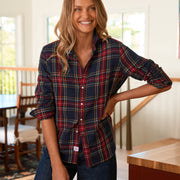 A woman modeling a long-sleeve button-up plaid shirt in a home interior setting. The shirt features a navy blue base with red, green, and yellow plaid stripes.