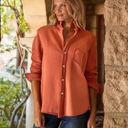 A woman wearing a rust-orange long-sleeve button-up shirt with a chest pocket, paired with dark blue jeans, standing in a rustic stone corridor.