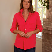 A woman wearing a bright red button-down shirt with rolled-up sleeves and dark blue jeans, standing in a home interior next to a brick column and window.