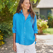 Woman wearing a blue shirt and white shorts standing outdoors with greenery in the background