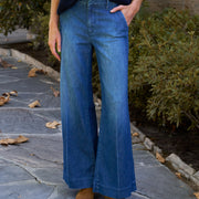A person wearing blue wide-leg denim jeans, a dark navy ribbed sweater, and tan ankle boots, standing on a stone patio with green shrubs in the background.