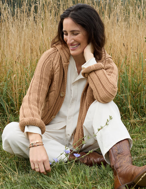 A woman sitting in a grassy field wearing a chunky tan cable-knit cardigan over a cream shirt and white pants, with brown cowboy boots., alternate view