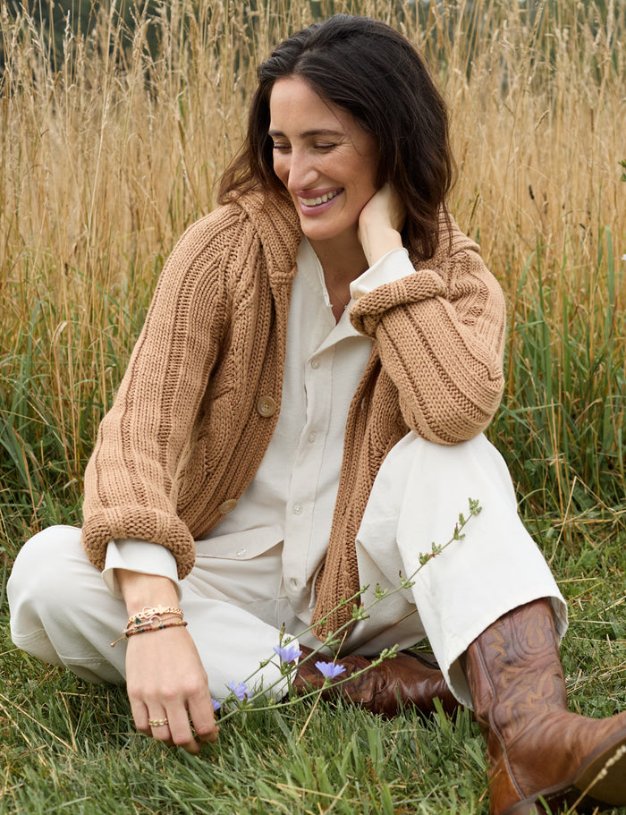 A woman sitting in a grassy field wearing a chunky tan cable-knit cardigan over a cream shirt and white pants, with brown cowboy boots.