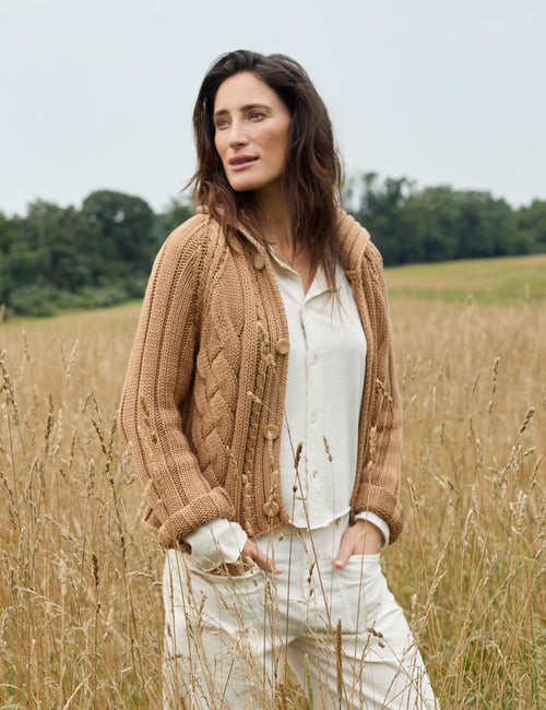 A woman standing in a field of dry grass wearing a tan cable-knit button-front cardigan over a white shirt and white pants., alternate view