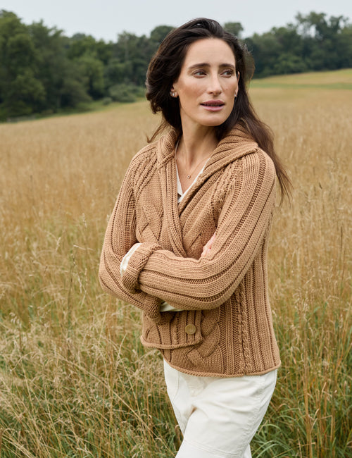 A woman stands in a field of dry golden grass wearing a chunky cable-knit brown hooded cardigan and white trousers., alternate view