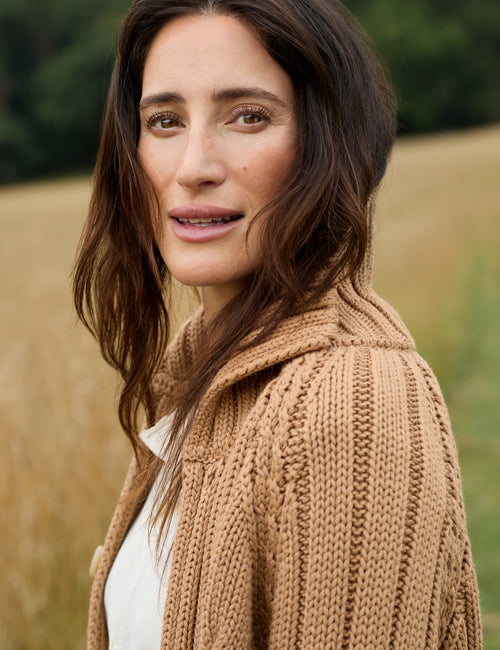 A woman with long brown hair wearing a chunky knit beige cardigan over a white top, photographed outdoors against a blurred field background., view 3