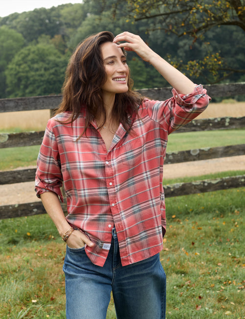 A woman wearing a red and gray plaid button-down shirt with rolled-up sleeves and blue jeans, standing outdoors in a grassy field with a wooden fence and trees in the background., alternate view