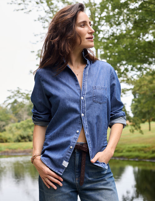 A woman wearing a blue denim button-up shirt and jeans stands outdoors near a lake with trees in the background., alternate view