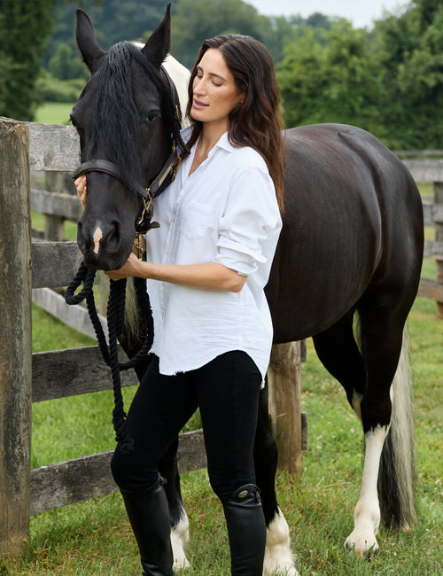 A woman standing outdoors next to a dark horse with white markings, wearing a white button-up shirt, black pants, and black riding boots, with a wooden fence and green trees in the background., alternate view