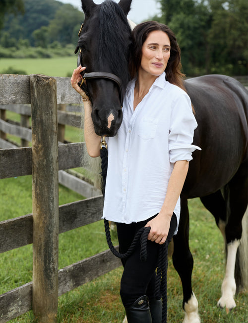 A woman standing outdoors beside a dark horse in a grassy field, wearing a white button-up shirt, black pants, and tall black riding boots, while holding the horse's lead rope., alternate view