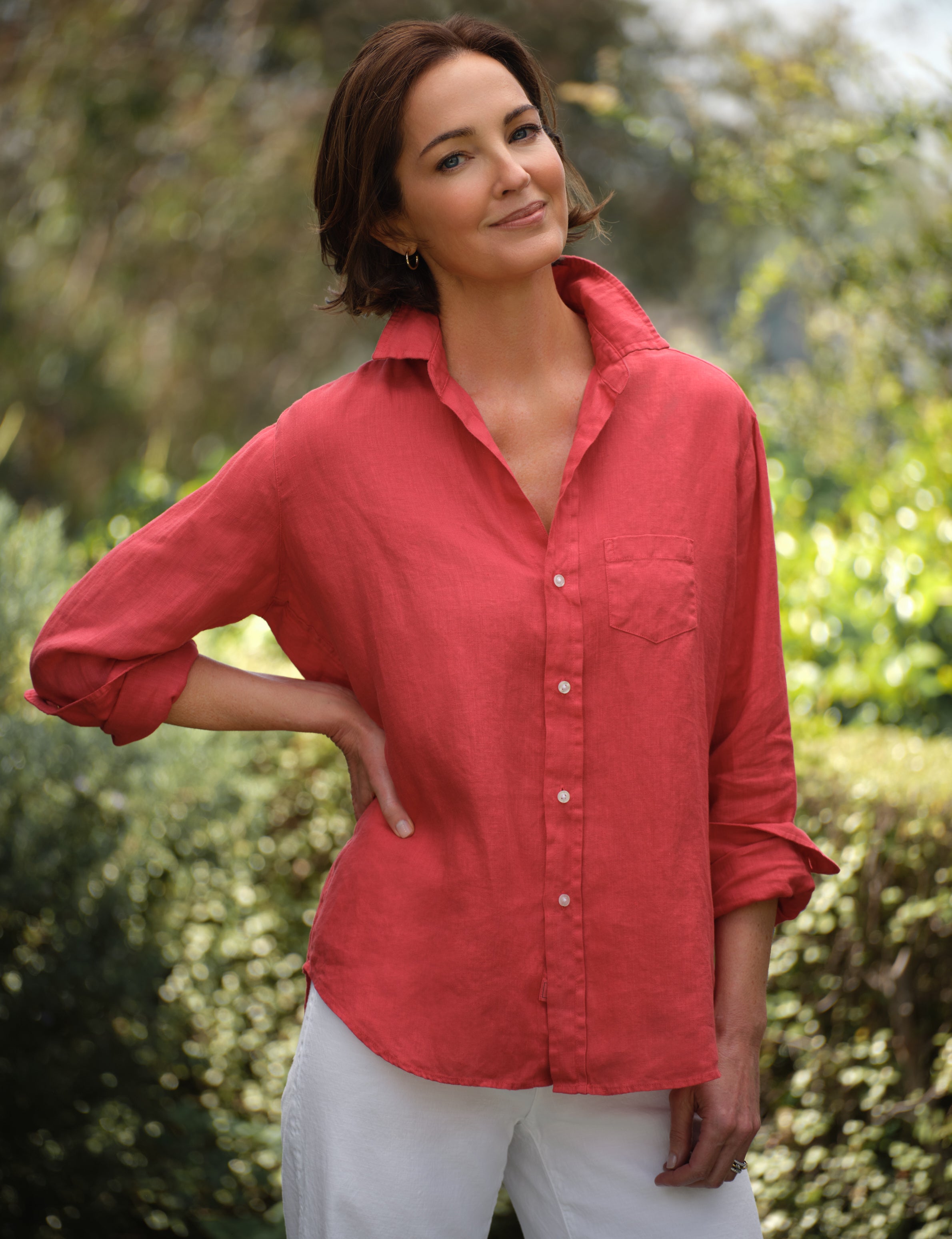 Woman wearing a red shirt and white pants standing outdoors with greenery in the background, front view
