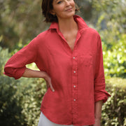 Woman wearing a red shirt and white pants standing outdoors with greenery in the background