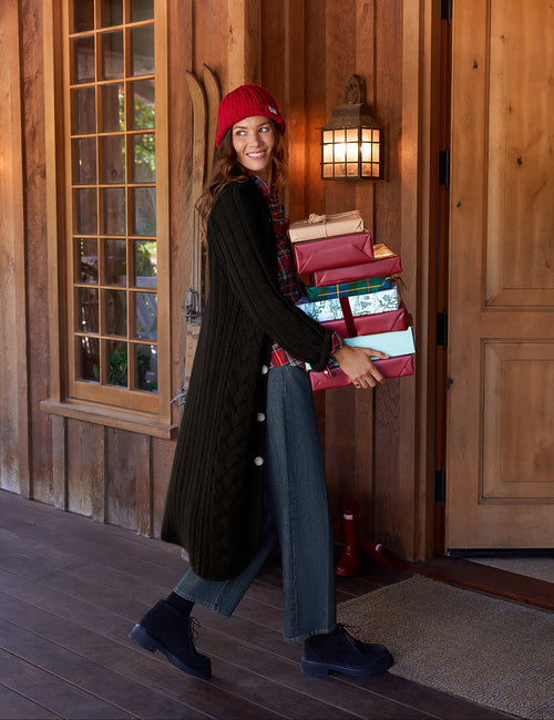 A woman wearing a black ribbed knit long coat, blue jeans, black boots, and a red knit beanie walks through a rustic wooden doorway while carrying a stack of colorful books., view 4
