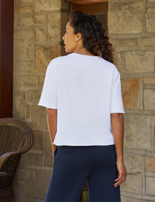A woman with curly dark hair is shown from the back, wearing a white short-sleeve crop top and navy blue pants, standing in front of a stone wall., alternate view