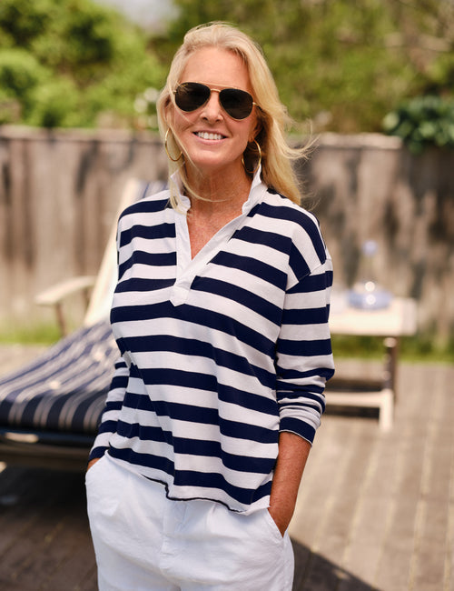 A woman wearing a navy and white horizontally striped long-sleeve polo shirt, white pants, and dark sunglasses, standing on a wooden deck with a patio chair and greenery in the background., alternate view