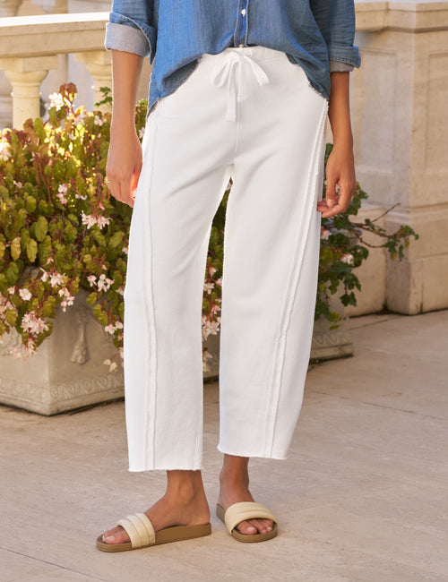 Person wearing white pants and a blue shirt standing outdoors with plants in the background, view 4