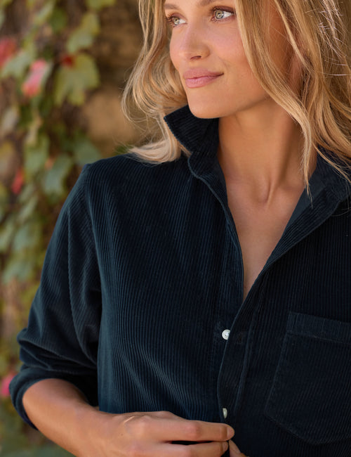 A close-up of a woman wearing a dark navy corduroy button-up dress, looking off-camera against a soft-focus background of green foliage., alternate view