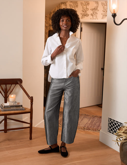 A woman stands in a warmly lit indoor room wearing a white button-up shirt, wide-leg metallic gray pants, and black flats. A wooden chair with books and a candle is visible on the left, and a doorway and wall sconce are in the background., view 4