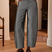 A person wearing gray barrel-leg cropped pants with a white button-up shirt and black flats, standing in a home interior with wood flooring.