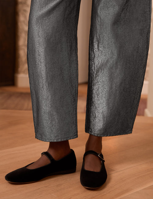 Close-up of a person wearing barrel-leg metallic gray pants and black flat Mary Jane shoes on a light wood floor., view 3