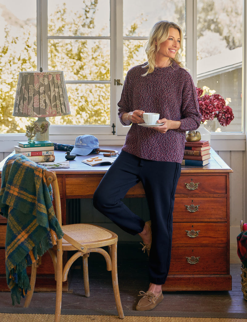 Woman in a cozy room with a desk, lamp, and books, holding a cup.