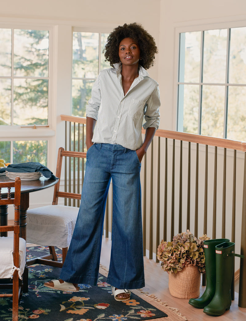 Woman standing in a room with large windows, wearing a green stripe shirt and blue jeans.