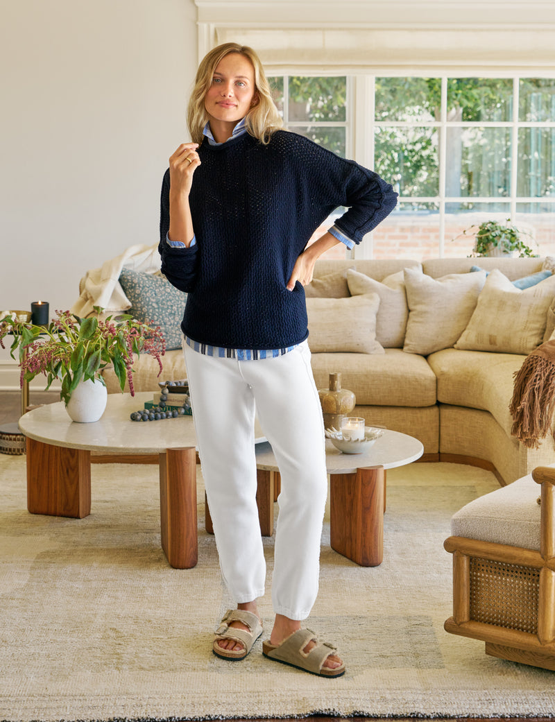 Woman standing in a living room wearing a navy sweater and white pants.