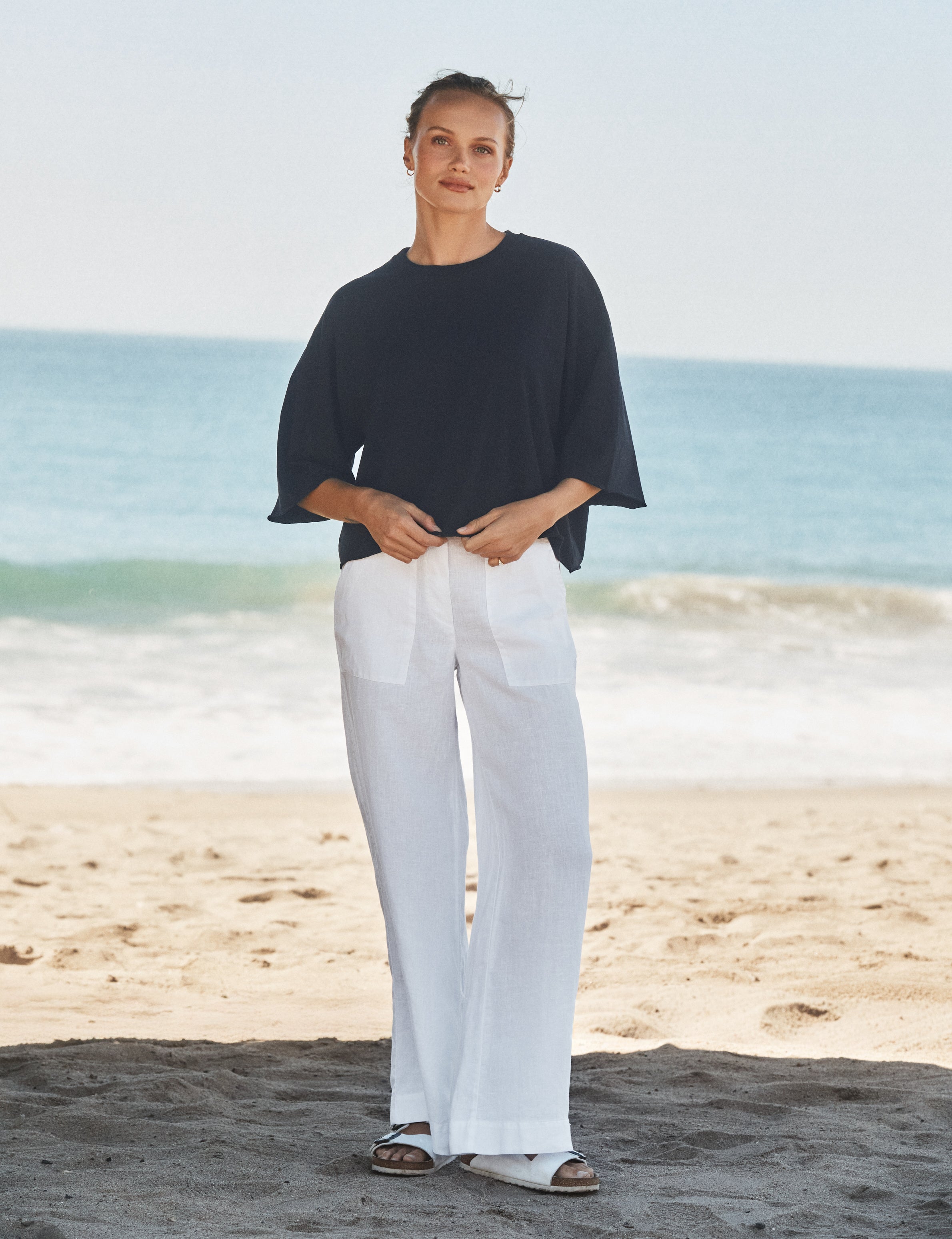 Woman wearing a navy top and white pants standing on a beach with ocean in the background, view 4
