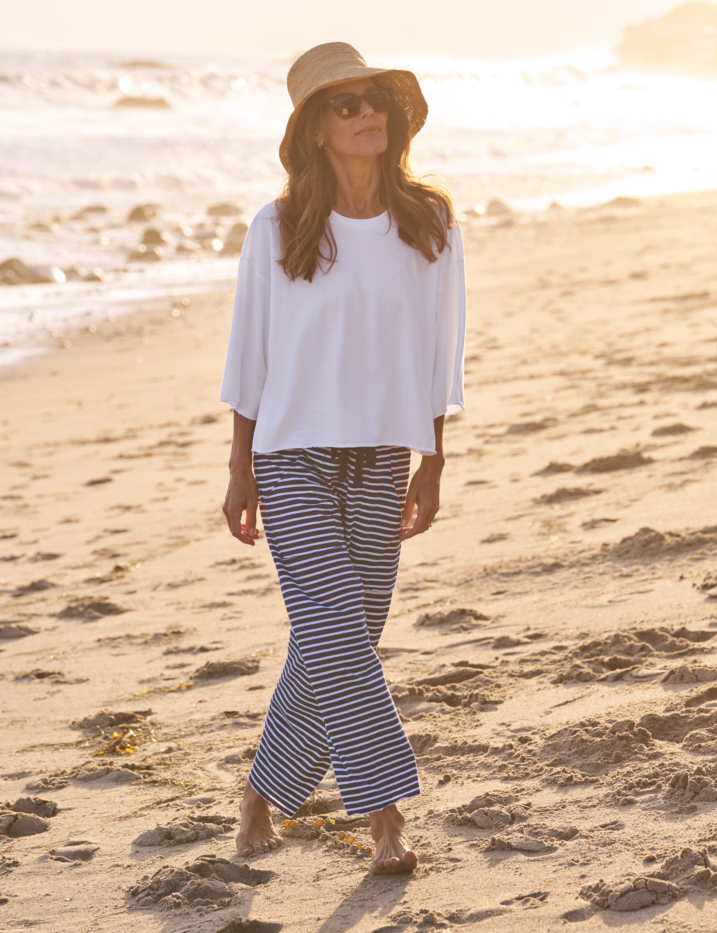 Woman in white top and striped pants standing on a sandy beach with sunset in the background, view 4