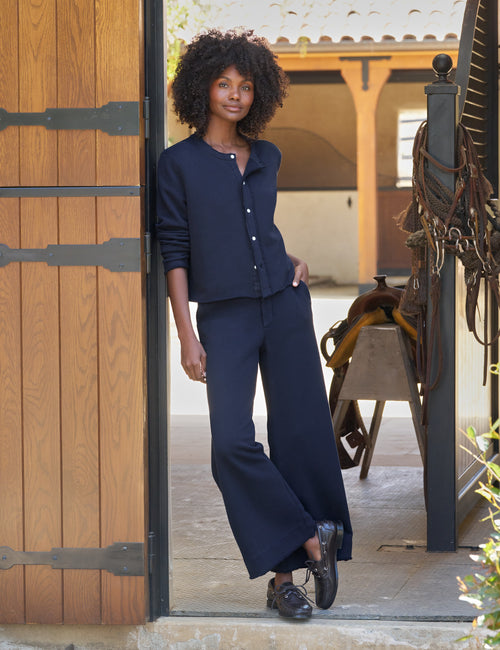 A woman with curly hair stands in a doorway wearing a navy blue long-sleeve cardigan and matching wide-leg pants, paired with black shoes. The setting appears to be a rustic indoor-outdoor space with wood paneling, a saddle, and warm architectural details in the background., front view