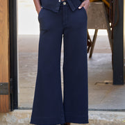 A person wearing dark navy wide-leg cropped pants with a matching navy button-front top and black lace-up shoes, standing in a doorway.