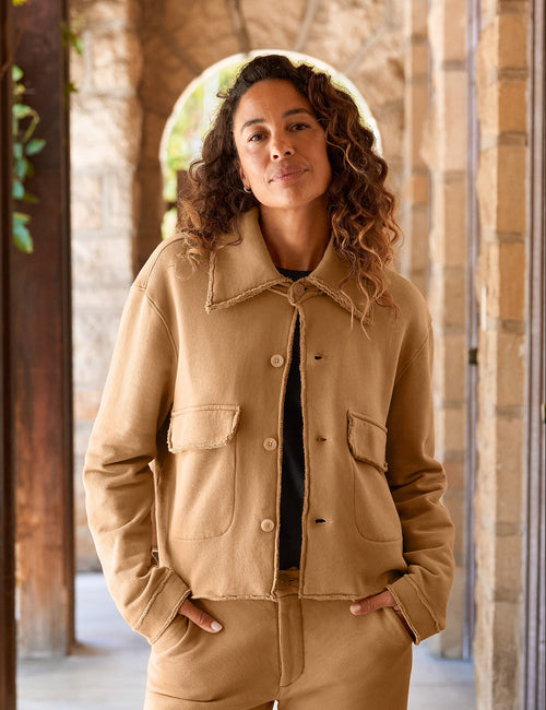 A woman stands in a stone and wood covered walkway wearing a camel-colored two-piece jacket and wide-leg pants set with a black top underneath., view 5
